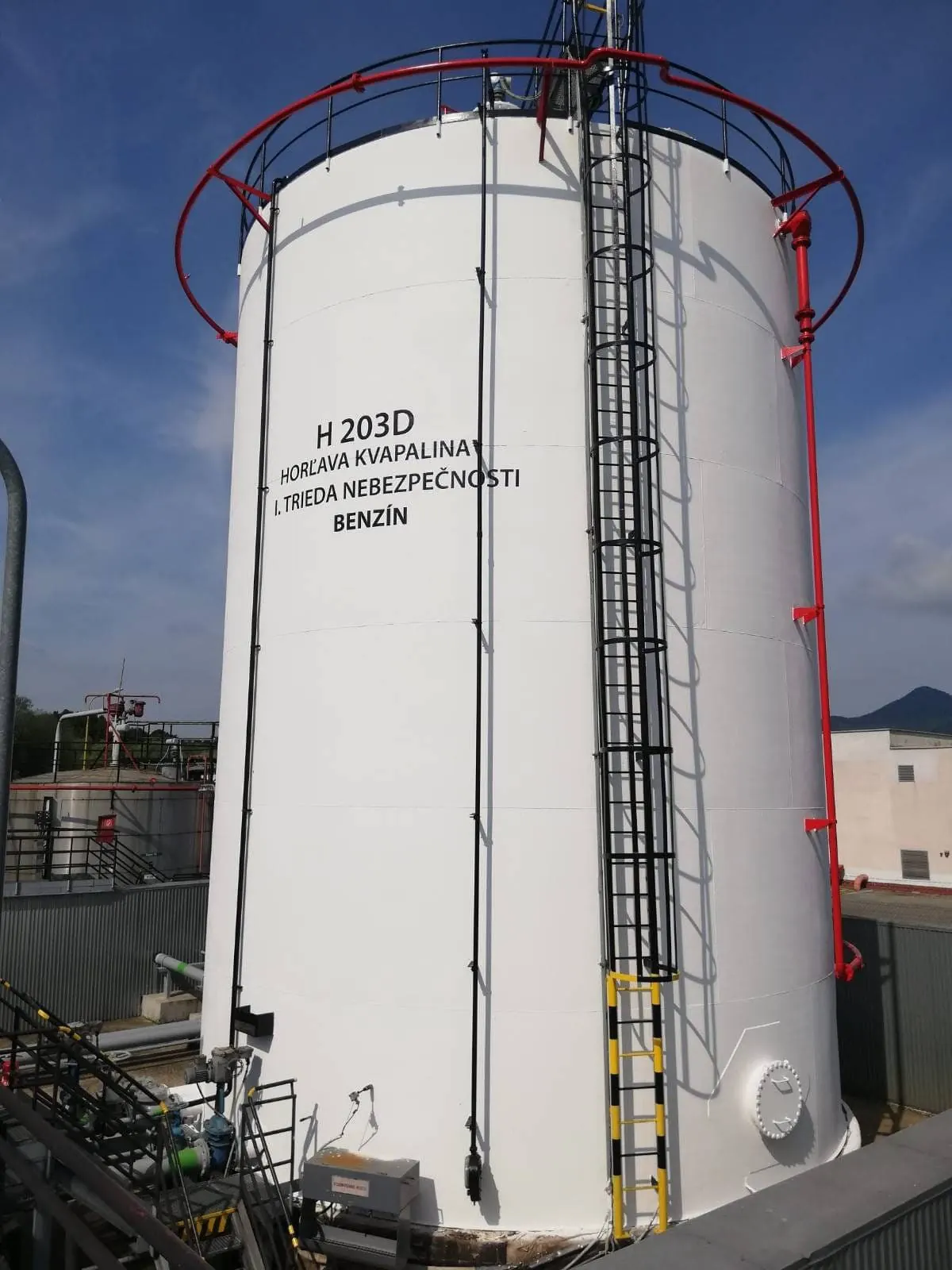 Weiß beschichteter Lagertank für Benzin mit Außenleiter und rotem Umlaufgeländer auf einem Tanklager.